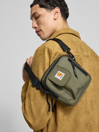 Männermodel trägt Essentials Bag mit Logo-Patch von Carhartt Work In Progress, olivgrünes Material, schwarze Akzente, Nahaufnahme Oberkörper. Geteiltes Hauptfach mit Reißverschluss, abnehmbarer Schulterriemen, Innenfach mit Reißverschluss, Logo-Patch sichtbar.