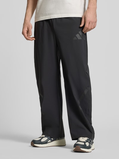 Männermodel trägt Oversized Jogpants mit Reißverschlusstaschen von adidas Sportswear in Schwarz, Elastischer-Material-Mix, Ganzkörperansicht von vorne.