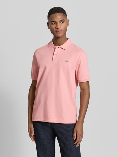 Męski model nosi Slim Fit Poloshirt mit Logo-Stitching von Gant z bawełny, krój slim fit, wyhaftowane logo, kołnierzyk polo, krótki rękaw, długość tyłu w rozmiarze M: 70 cm, jasnoróżowa koszulka na tle neutralnym, całkowite ujęcie od przodu.