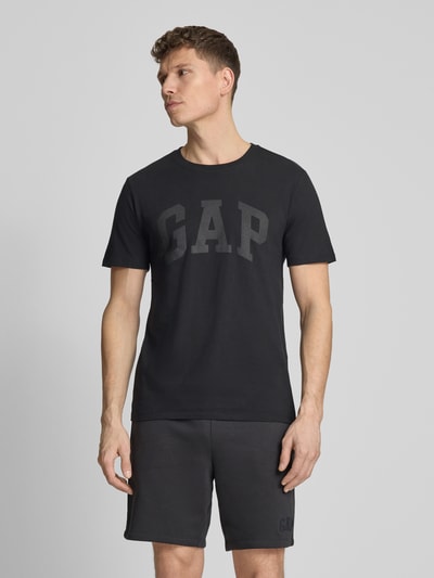 Männermodel trägt schwarzes Herren T-Shirt mit Logo und Rundhalsausschnitt von GAP aus reiner Baumwolle, Regular-Fit, Ganzkörperansicht von vorne. Rückenlänge bei Größe M: 72 cm.