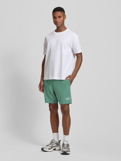 Männermodel trägt Shorts mit elastischem Bund und Logo von EA7 Emporio Armani in Grün, Regular Fit, Eingrifftaschen, Ganzkörperansicht von vorne.