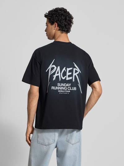 Männermodel trägt T-Shirt mit Statement-Print Modell 'SRC® Pacer' von ANOTHER COTTON LAB in Schwarz, lockerer Boxy Fit, reine Baumwolle, Rückenlänge 69 cm, Ganzkörperansicht Rückansicht