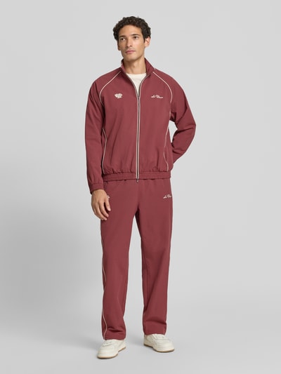 Männermodel trägt Trackpants mit Logo-Stitching Modell 'Seersucker' von Les Deux in Rot mit Strukturmuster, Relaxed Fit, Ganzkörperansicht von vorne.