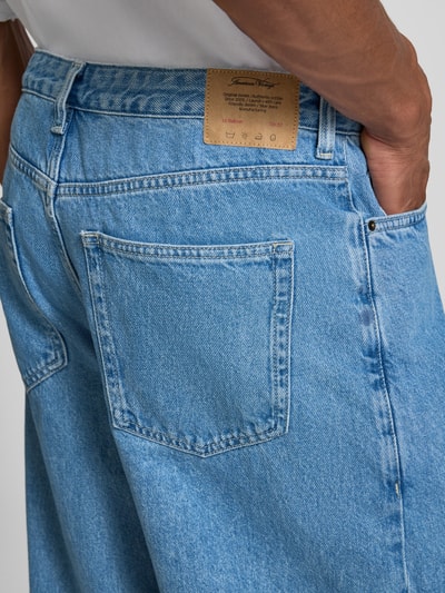 Mężczyzna nosi jeansy z wpuszczanymi kieszeniami Jeans mit Eingrifftaschen Modell 'JOYBIRD' von American Vintage, krój relaxed fit, jasnoniebieski denim, widoczna metka, kontekst: szerokość w rozmiarze 32/32: 40 cm, długość wewnętrzna nogawki: 74 cm, zapięcie na guzik i zamek błyskawiczny, detaliczne ujęcie tylnej kieszeni, zbliżenie.