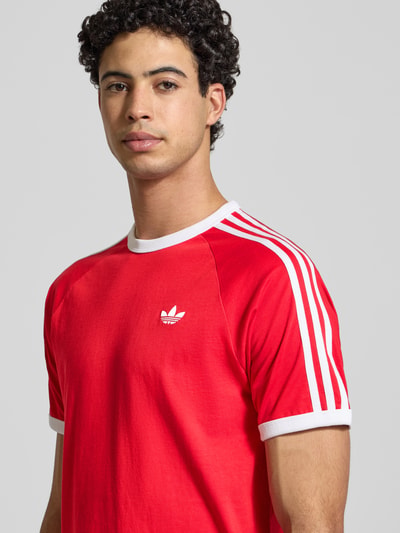 Männermodel trägt T-Shirt mit Label-Stitching von adidas Originals in Rot mit weißen Streifen, reine Baumwolle, Regular-Fit, Nahaufnahme Oberkörper.