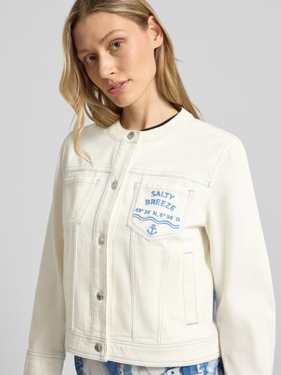 Frauenmodel trägt Jeansjacke mit Motiv- und Statement-Stitching von Marc Cain in Offwhite mit blauen Nähten aus Baumwoll-Mix mit Stretch-Anteil, gerader Schnitt, Rundhalsausschnitt, Knopfleiste, Brusttaschen und seitlichen Eingrifftaschen; Rückenlänge 50 cm, Schulterbreite 42 cm, Brustweite 46 cm, Ärmellänge 49 cm; Nahaufnahme Oberkörper.