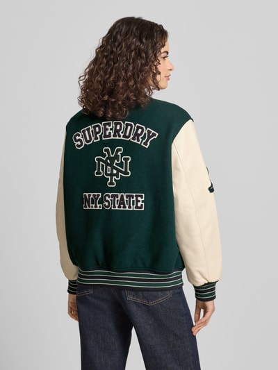 Frauenmodel trägt College Jacke mit Motiv- und Logo-Stitchings von Superdry in Dunkelgrün mit Woll-Mix, Lockerer-Schnitt, Stehkragen, Druckknopfverschluss, elastische Abschlüsse, Rückenlänge 62 cm, Schulterbreite 49 cm, Brustweite 60 cm, Ärmellänge 60 cm, Ganzkörperansicht Rückansicht.