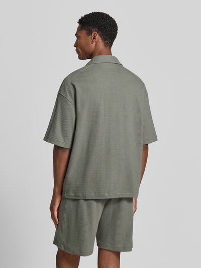 Męski model nosi Relaxed Fit Freizeithemd mit Strukturmuster od Drykorn z bawełny, kołnierz z połami, krótki rękaw, listwa guzikowa, kolor khaki, fakturowany wzór, długość tyłu w rozmiarze M: 69 cm, szerokość w rozmiarze M: 60 cm, długość rękawów w rozmiarze M: 26 cm, widok całej sylwetki z tyłu.