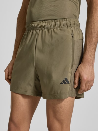 Männermodel trägt Shorts mit elastischem Bund von adidas Training in Khaki mit Label-Print und Reißverschlusstaschen, Nahaufnahme Oberschenkel. Lockere Passform, Bundweite 38 cm, Innenbeinlänge 14 cm.
