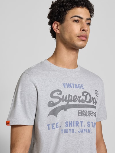Męski model nosi Relaxed Fit T-Shirt mit Logo-Stitching Modell 'Heritage' von Superdry, mieszanka bawełny, okrągły dekolt, długość tyłu w rozmiarze M: 71 cm, jasnoszary, nadruk logo, Nahaufnahme górnej części ciała.