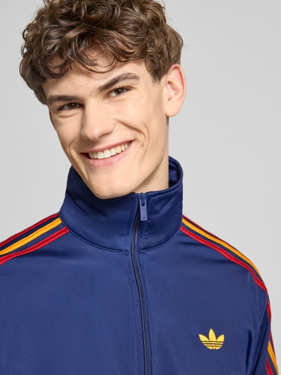 Männermodel trägt Regular Fit Sweatjacke mit Logo-Stitching Modell 'FIREBIRD TT' von adidas Originals in Blau mit Kontraststreifen, Nahaufnahme Oberkörper.