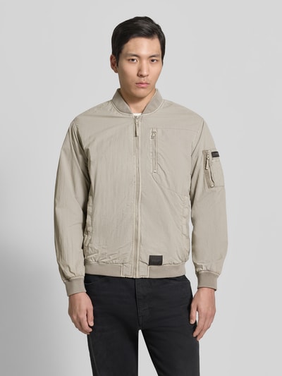 Männermodel trägt Blouson mit Label-Patches Modell 'IBE' von khujo in Beige, Gerader-Schnitt, Stehkragen, Reißverschluss, Ganzkörperansicht von vorne. Brusttasche mit Reißverschluss, Reißverschlusstasche am Ärmel, seitliche Eingrifftaschen, Rückenlänge 64 cm, Schulterbreite 48 cm, Brustweite 56 cm, Ärmellänge 62 cm.