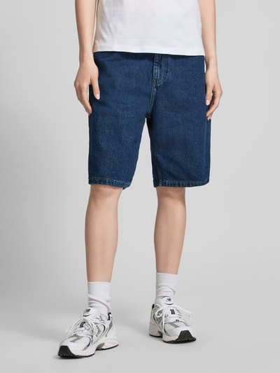 Frauenmodel trägt Relaxed Fit Jeans Shorts aus reiner Baumwolle Modell 'Brandon' von Carhartt Work In Progress in Dunkelblau, 5-Pocket-Design, Knopf-Reißverschluss, Label-Patch, Ganzkörperansicht von vorne, Bundweite 41 cm, Innenbeinlänge 24 cm bei Größe S.