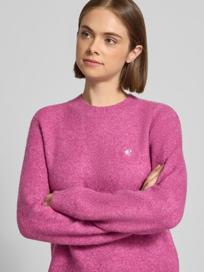 Vrouwenmodel draagt gebreide pullover met labeldetail Model 'Curtis' van Carhartt Work In Progress, materiaalmix met wol, getailleerde pasvorm in roze, ribboorden en ronde hals, ruglengte maat S 55 cm, mouwlengte 60 cm, detailopname bovenlichaam.