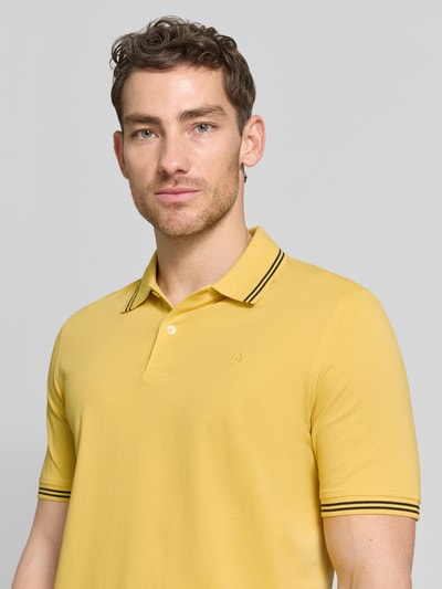 Männermodel trägt Regular Fit Poloshirt mit Logo-Stitching von bugatti, gelb, Reine Baumwolle, Polokragen, kurze Knopfleiste, Label-Detail, Nahaufnahme Oberkörper.