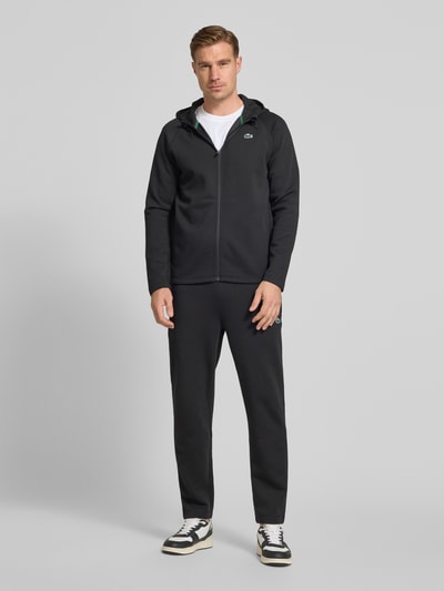 Męski model nosi Regular Fit Sweatjacke mit Baumwoll-Anteil od Lacoste, efekt: jednolity kolor, krój regular fit, zamek błyskawiczny, naszywka z logo, kaptur z tunel, długi rękaw, wpuszczane kieszenie, scuba, mieszanka bawełny, ganzkörperansicht z przodu.