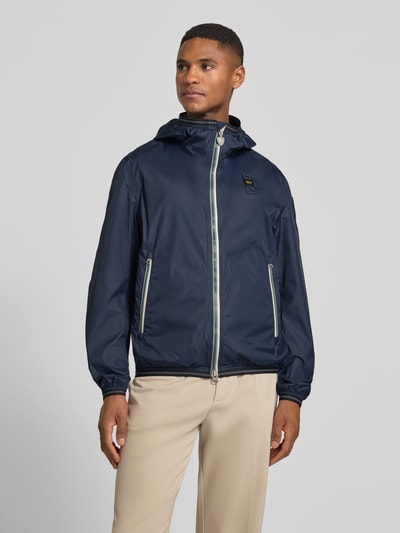 Männermodel trägt Jacke mit Label-Patches Modell 'Dodge' von Blauer Usa in Dunkelblau mit Kapuze, Zweiwege-Reißverschluss, Seitliche Reißverschlusstaschen, elastische Abschlüsse, Gerader-Schnitt, Ganzkörperansicht von vorne.