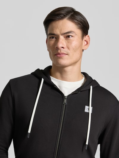 Männermodel trägt Sweatjacke mit Logo-Patch und Känguru-Tasche von Scotch & Soda aus reiner Baumwolle in Schwarz, Nahaufnahme Oberkörper.