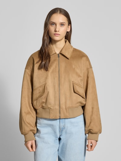 Modelka kobieca ma na sobie Regular Fit Bomberjacke mit Umlegekragen od Guess Jeans, beżowy kolor, zamek, detal kieszeni, krój regular fit, długość tyłu w rozmiarze S: 47 cm, szerokość ramion w rozmiarze S: 56 cm, szerokość klatki piersiowej w rozmiarze S: 45 cm, długość rękawa w rozmiarze S: 40 cm, widok całej sylwetki z przodu.