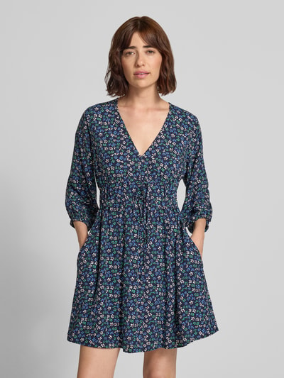 Frauenmodel trägt Minikleid aus reiner Viskose mit floralem Allover-Print von GAP in dunklem Blau mit bunten Blumen, taillierte Passform, V-Ausschnitt, 3/4-Arm, fließende Textur, Ganzkörperansicht von vorne.