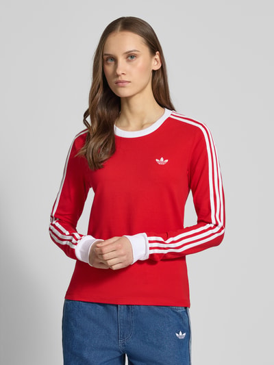 Frauenmodel trägt Longsleeve mit Rundhalsausschnitt und Logo-Stitching von adidas Originals, roter Baumwoll-Mix mit Stretch-Anteil, körpernaher Schnitt, weiße Streifen und Details, Rückenlänge bei Größe S: 53 cm, Nahaufnahme Oberkörper.