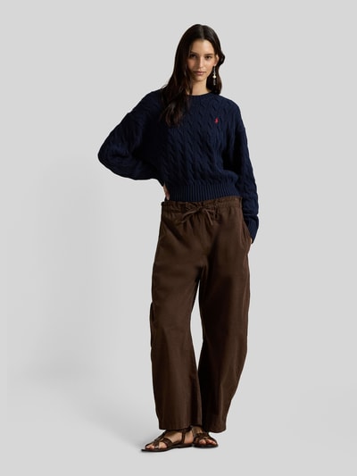 Modelka nosi Regular Fit Strickpullover aus reiner Baumwolle od Polo Ralph Lauren, granatowy sweter z bawełny, splot warkoczowy, logo, krój regular fit, okrągły dekolt, długi rękaw, mankiety i dół prążkowane, widok całej sylwetki z przodu.
