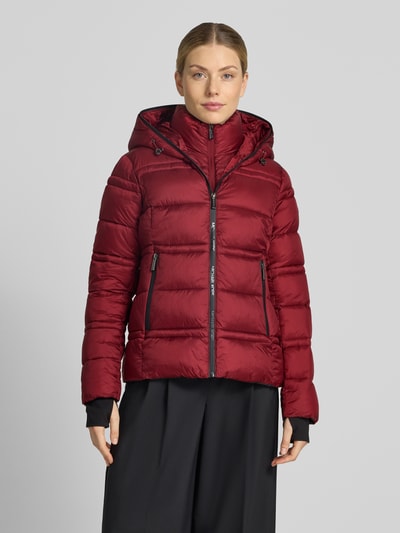 Frauenmodel trägt Steppjacke mit Kapuze und Reißverschlusstaschen von MICHAEL Michael Kors in Weinrot, sichtbare Steppnähte, glatte Textur, Ganzkörperansicht von vorne.