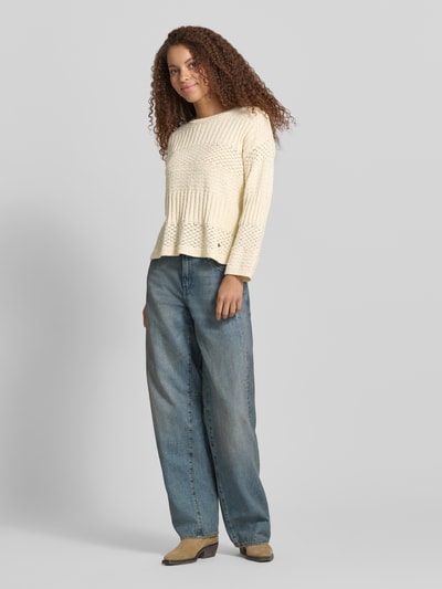 Frauenmodel trägt Boxy Fit Strickpullover aus reiner Baumwolle Modell 'ALAYA' von Pepe Jeans in cremefarbener Strickoptik mit Strukturmuster, Rundhalsausschnitt, langen Ärmeln und asymmetrischem Saum in Ganzkörperansicht von vorne.