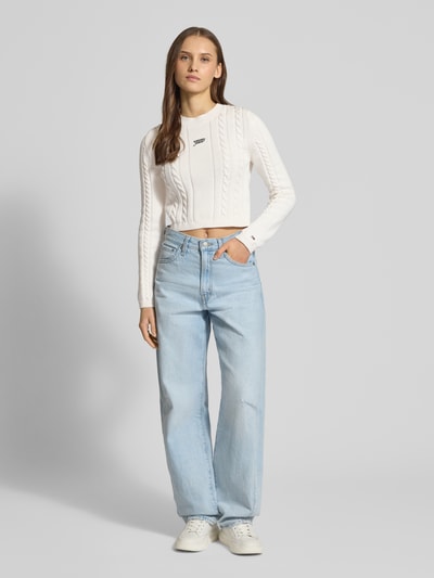 Frauenmodel trägt Slim Fit Strickpullover aus reiner Baumwolle von Tommy Jeans in Optik: Unifarben, Strickoptik, mit Logo-Detail und Label-Schriftzug, Rundhalsausschnitt, Langarm, gerippte Abschlüsse, Materialverarbeitung: Strick, Ganzkörperansicht von vorne.