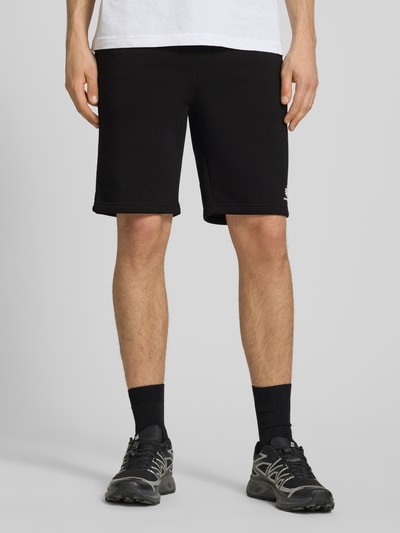 Männermodel trägt schwarze Shorts mit Eingrifftaschen und Logo-Print von Alpha Industries aus Baumwoll-Mix, Relaxed-Fit, Ganzkörperansicht von vorne. Bundweite Größe M 39 cm, Innenbeinlänge 24 cm, elastischer Bund mit Kordelzug, Gesäßtasche.