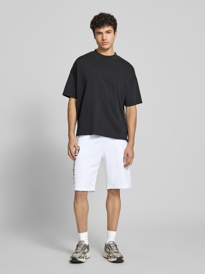 Männermodel trägt Shorts mit Logo-Print und -Stitching von CHAMPION in Weiß, lockerer Schnitt, elastischer Bund, Ganzkörperansicht von vorne.