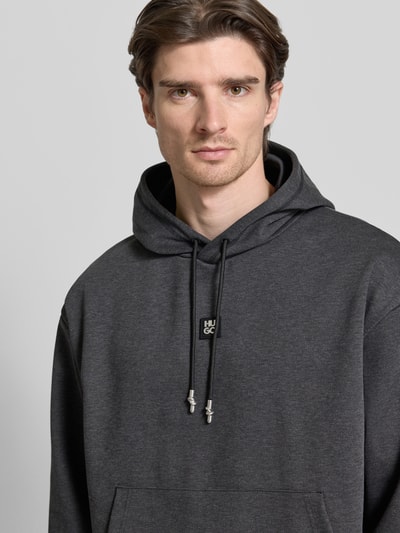 Mężczyznę model nosi Regular Fit Hoodie aus Baumwoll-Mix Modell 'DINEO' von HUGO, ciemnoszary, jednolity, z kapturem, label patch, metaliczne akcenty, kieszeń kangurka, tekstura sweat, krój regular fit, widok z przodu, zbliżenie na górną część ciała.