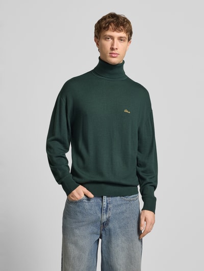 Männermodel trägt Loose Fit Rollkragenpullover aus Woll-Mix von BLKVIS in Dunkelgrün mit gerippten Abschlüssen und Label-Stitching, Nahaufnahme Oberkörper. Rückenlänge bei Größe M: 68 cm, Ärmellänge: 57 cm.