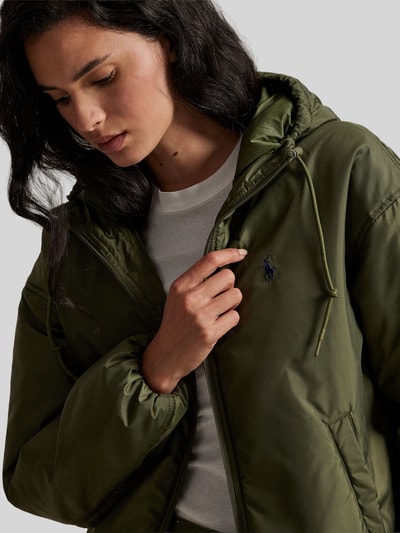 Kobiece modelka nosi Relaxed Fit Blouson mit Zweiwege-Reißverschluss od Polo Ralph Lauren, jednolity kolor oliwkowy, logo na piersi, kaptur z troczkiem, elastyczne mankiety i dół, boczne kieszenie; zbliżenie na górną część ciała.