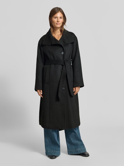 Frauenmodel trägt Mantel mit Baumwoll-Anteil von MYON in Schwarz mit Reverskragen, Taillienband und Eingrifftaschen, Ganzkörperansicht von vorne.
