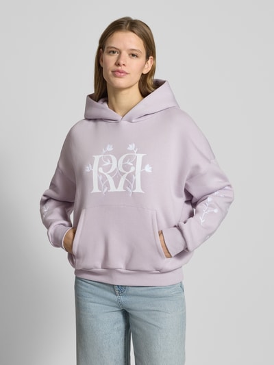 Frauenmodel trägt Relaxed Fit Hoodie mit Logo-Print von Review in Flieder mit floraler Stickerei, Känguru-Tasche, Ganzkörperansicht von vorne.