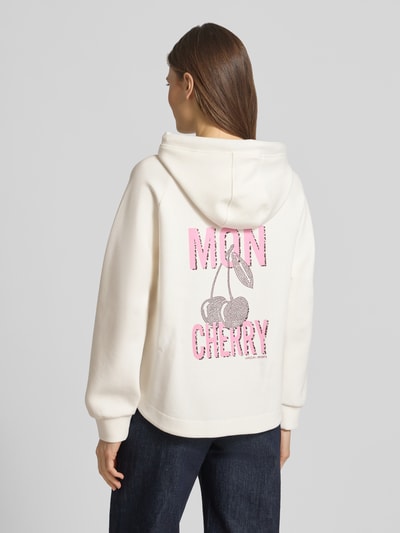 Modelka nosi Relaxed Fit Hoodie mit Motiv-Print od Marc Cain z mieszanki bawełny, krój relaxed fit, nadruk z napisem i motywem, kaptur z tunelem, elastyczne zakończenia rękawów, jasny kolor, wyraźna tekstura dzianiny, widoczny nadruk z tyłu, widoczne detale szwów; zdjęcie w ujęciu całopostaciowym z tyłu.