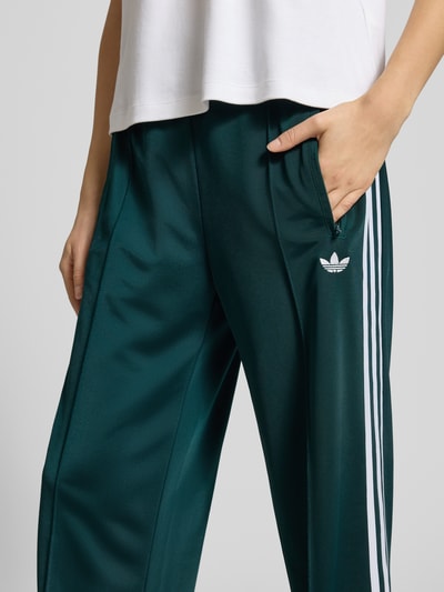 Frauenmodel trägt Sweatpants mit elastischem Bund von adidas Originals in Dunkelgrün mit Label-Stitching und Reißverschlusstaschen, Nahaufnahme Oberkörper, lockerer Schnitt, Innenbeinlänge bei Größe S: 80 cm.