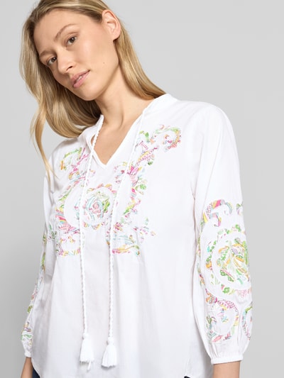 Frauenmodel trägt Bluse mit Schlüsselloch-Ausschnitt und Schnürung von Emily Van den Bergh aus Reine Baumwolle, Straight Fit, weiß mit buntem Muster, 3/4-Arm, elastische Ärmelabschlüsse, abgerundeter Saum, Ganzkörperansicht von vorne.