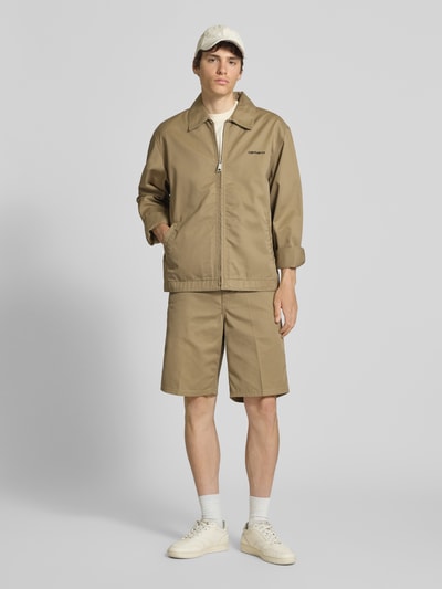 Męski model nosi Jacke mit Label-Stitching und Eingrifftaschen od Carhartt Work In Progress, beżowa, krój regular fit, wykładany kołnierz, zamek błyskawiczny, wyhaftowane logo, wpuszczane kieszenie, długość tyłu w rozmiarze M: 68 cm, szerokość ramion w rozmiarze M: 59 cm, szerokość klatki piersiowej w rozmiarze M: 60 cm, długość rękawów w rozmiarze M: 20 cm, widok całej sylwetki z przodu.