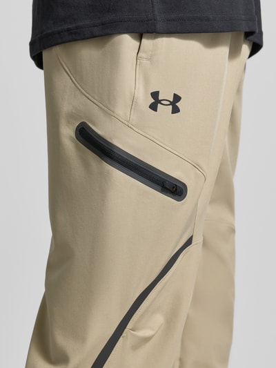 Männermodel trägt Regular Fit Sweatpants mit Logo-Print Modell 'Unstoppable' von Under Armour, Beige mit Cargo-Tasche und Reißverschluss, Elastischer-Material-Mix, Nahaufnahme Oberschenkel.
