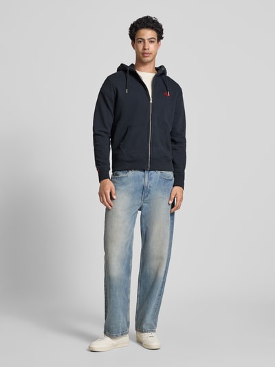 Männermodel trägt Sweatjacke mit Kapuze und Logo-Stitching von Superdry, dunkles Blau, Baumwoll-Mix, Regular-Fit, Ganzkörperansicht von vorne, seitliche Eingrifftaschen, Kordelzug, Reißverschluss, gerippte Abschlüsse, Label-Detail, Rückenlänge 66 cm, Ärmellänge 62 cm.