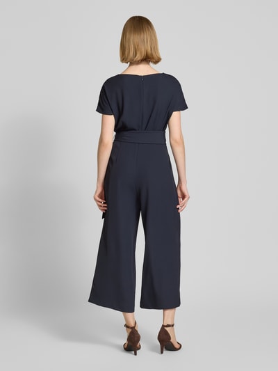 Vrouwenmodel draagt Jumpsuit mit Stretch-Anteil van Betty Barclay in donkerblauw, regular fit, korte mouwen, riem in taille, 7/8-lengte, coupenaden, detail op de rug, ruglengte 38 cm, borstwijdte 37 cm, mouwlengte 23 cm, totale lengte 127 cm, binnenbeenlengte 58 cm. Volledige lichaamsweergave achteraan.