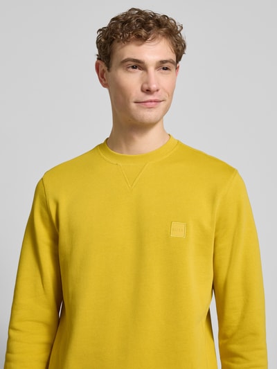 Männermodel trägt Relaxed Fit Sweatshirt aus reiner Baumwolle Modell 'WESTART' von BOSS Orange in Unifarbe Gelb, Rundhals, Label Patch, Nahaufnahme Oberkörper.