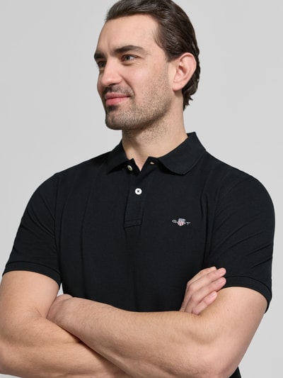 Męski model nosi Slim Fit Poloshirt mit Logo-Stitching von Gant, czarna bawełna, krój slim fit, logo, polo kołnierzyk, krótki rękaw, półzbliżenie górnej części ciała