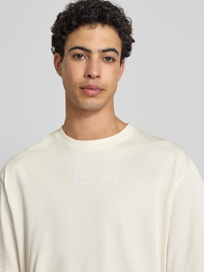 Männermodel trägt T-Shirt mit Label-Stitching und Rundhalsausschnitt von KARL KANI, cremefarbene Reine Baumwolle, Relaxed-Fit, Nahaufnahme Oberkörper. Rückenlänge bei Größe M: 70 cm.