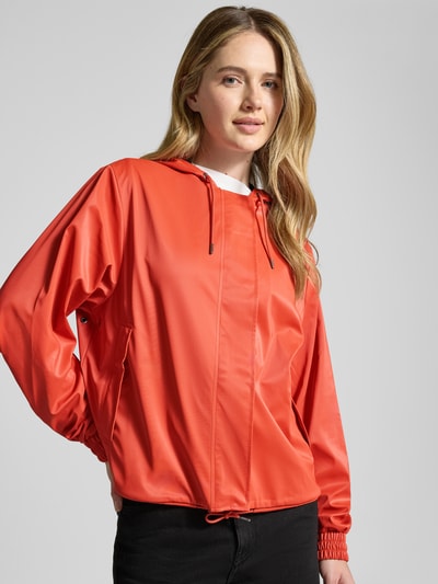 Modelka nosi Jacke mit Abdeckleiste od RAINS w kolorze czerwonym, prosta forma, kaptur ze ściągaczem, długi zamek z listwą, ściągacz w talii, elastyczne mankiety, długość pleców w rozmiarze S 58 cm, szerokość ramion w rozmiarze S 43 cm, szerokość w klatce piersiowej w rozmiarze S 55 cm, długość rękawa w rozmiarze S 63 cm, matowa faktura, widok z przodu, zbliżenie na górną część ciała.