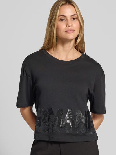 Vrouwenmodel draagt T-shirt met paillettenbesatz van Armani Exchange in zwart, puur katoen, los model, ronde hals, labelprint, ruglengte maat S 45 cm, close-up bovenlichaam.