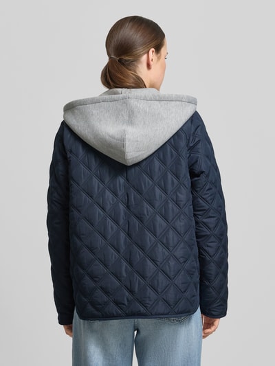 Kobiece modelka nosi Regular Fit kurtka z efektem pikowania z kapturem model 'Fenya' od Soyaconcept, wpuszczane kieszenie, listwa guzikowa, kolor granatowy, długość tyłu 65 cm, szerokość ramion 40 cm, szerokość w rozmiarze S 54 cm, długość rękawów 65 cm, widok całej sylwetki z tyłu.