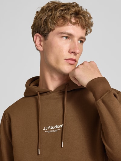 Männermodel trägt Regular Fit Hoodie aus Baumwoll-Mix Modell 'Soho' von JACK & JONES in Braun mit Logoprint, Nahaufnahme Oberkörper. Baumwoll-Mix, Kapuze mit Tunnelzug, Känguru-Tasche, elastische Abschlüsse, Rückenlänge 66 cm, Ärmellänge 65 cm.